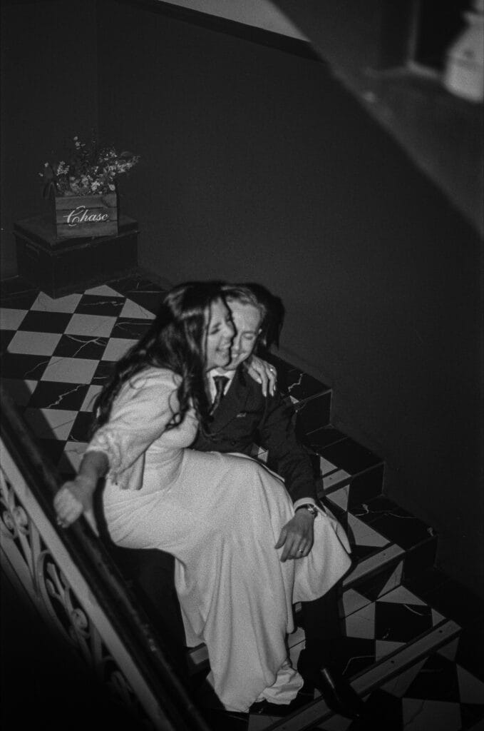 A woman in a white dress and a man in a suit sit closely together on a checkered staircase, with a flower arrangement visible in the background.