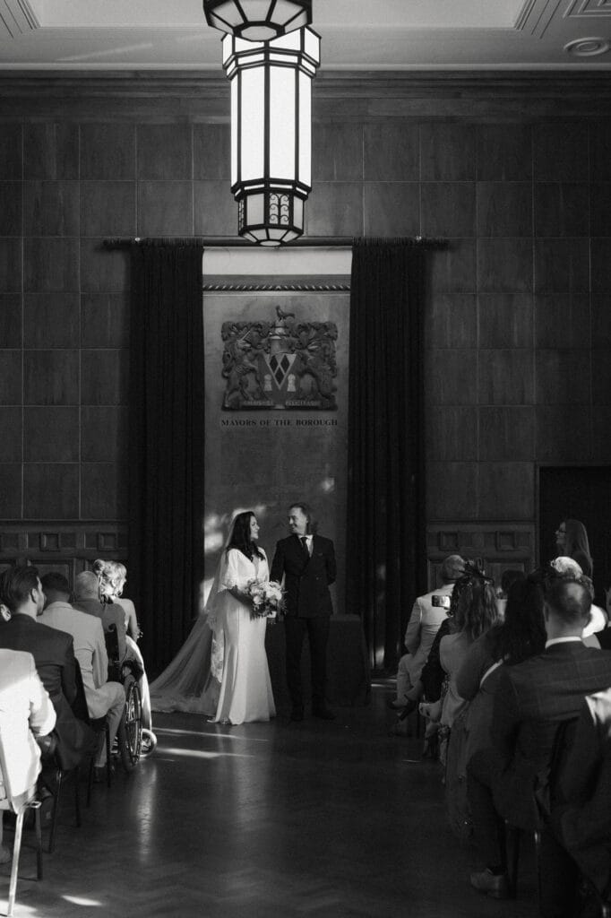 A bride and groom stand together at the front of a formal room, facing seated guests during a wedding ceremony.