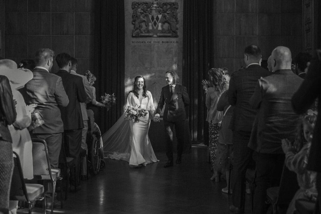 A bride and groom walk down the aisle together as guests stand and applaud in a formal indoor setting.