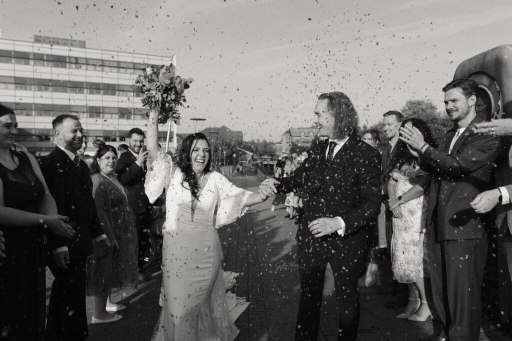 A newlywed couple walks outside, holding hands and smiling, as guests on both sides throw confetti in celebration.