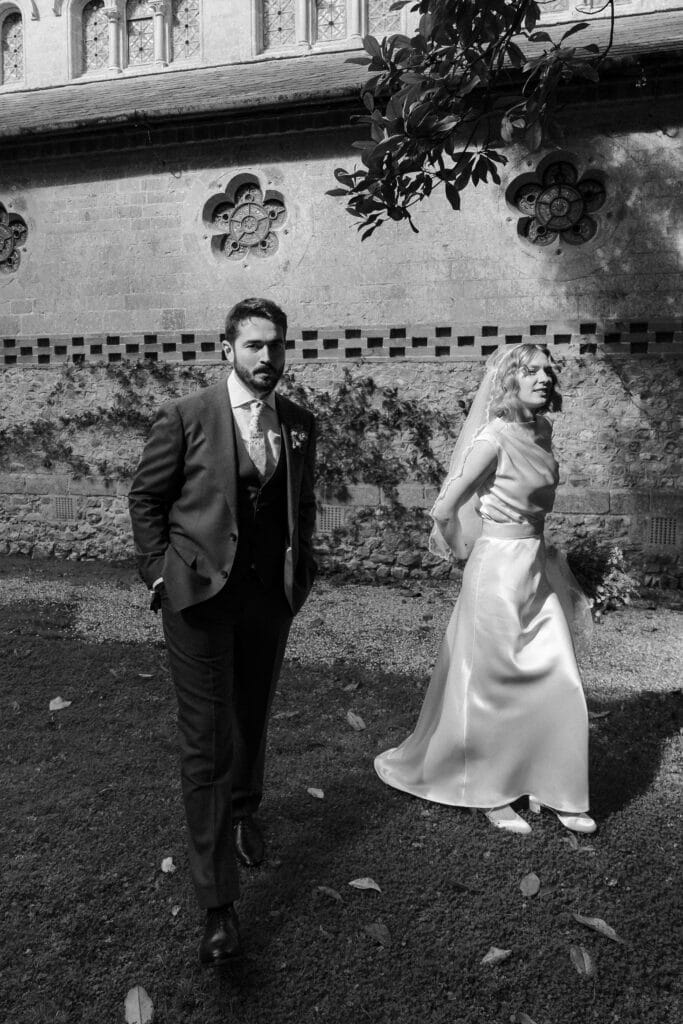 A bride in a satin dress and veil walks beside a groom in a suit outside a stone building with decorative windows.