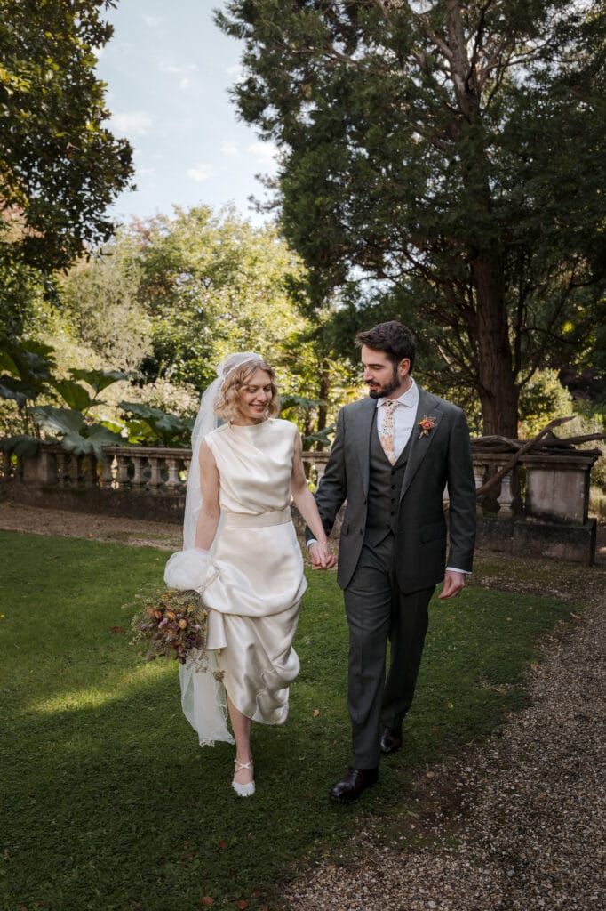 A bride in a white dress and a groom in a dark suit walk hand-in-hand on a garden path, surrounded by greenery.