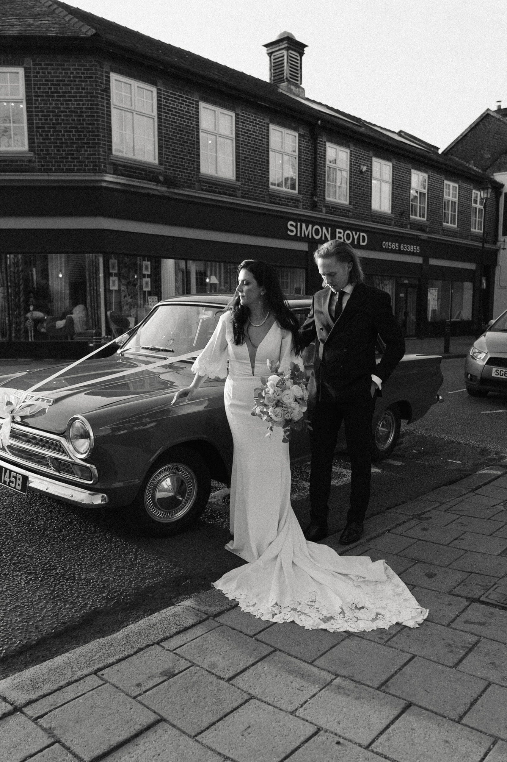 0336 CE BW Lost Found Knutsford © LOUMAY All Rights Reserved PLMP6610 scaled A bride in a white dress and a groom in a suit stand beside a vintage car on a street in front of a shop called Simon Boyd.