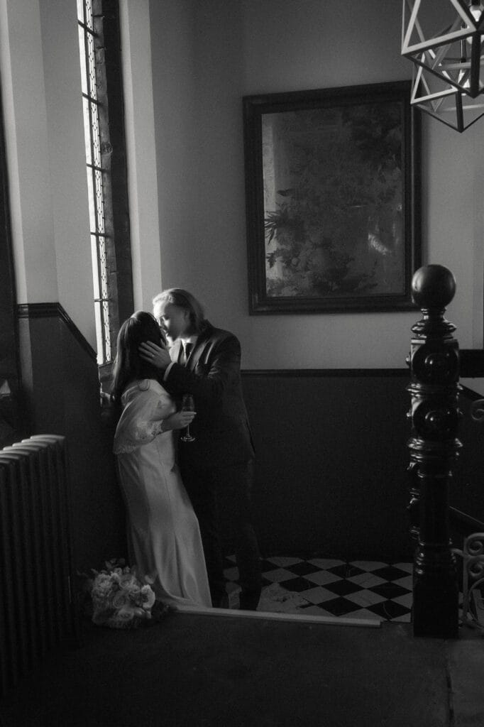 A couple in formal attire stands close by a window in a dimly lit hallway with black-and-white checkered floor tiles; a bouquet lies on the floor nearby.