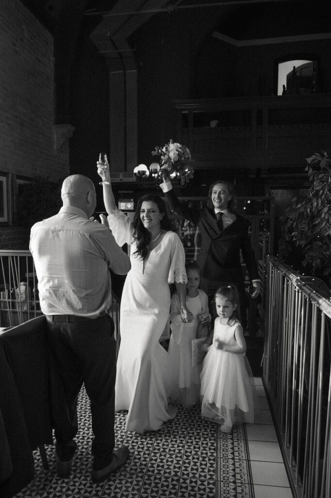 A couple in wedding attire enters a venue with two young girls; a man stands nearby, facing them, as the group smiles and celebrates.