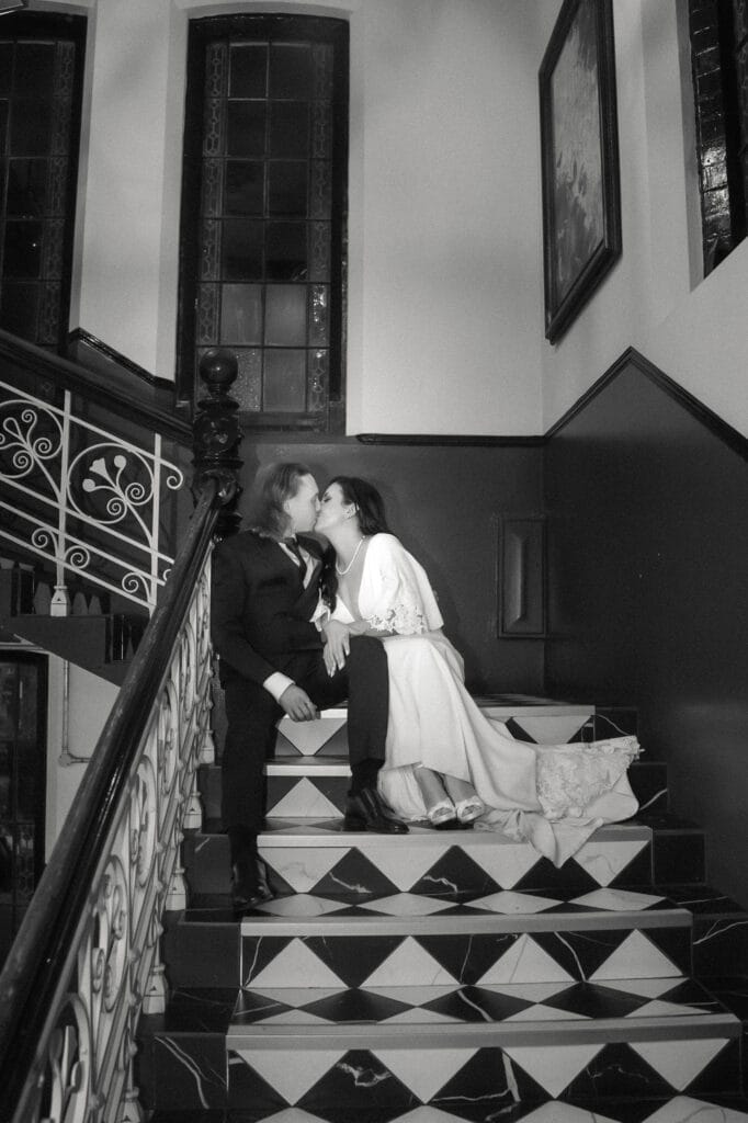 Two people in formal attire sit on a staircase, kissing. One wears a suit and the other a long dress. The scene is indoors with patterned steps and decorative railings.