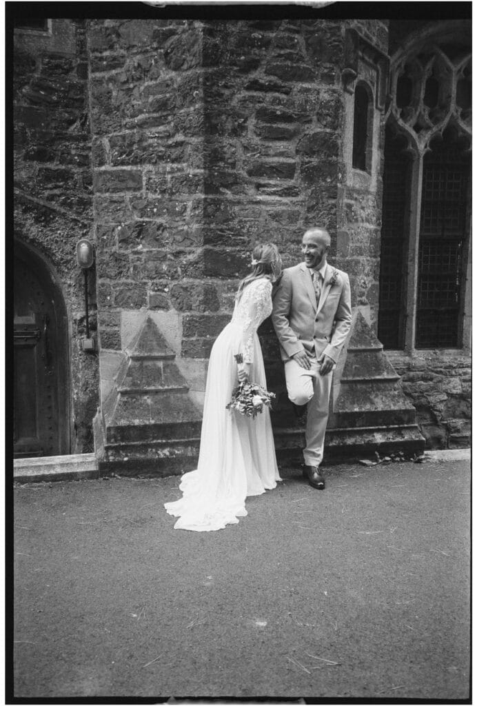 A bride in a long-sleeved gown leans toward a groom in a light suit as they stand together outside a stone building.