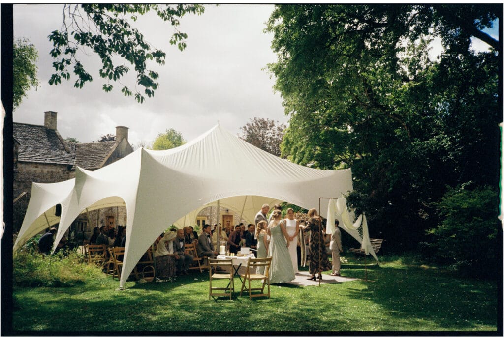 A group of people gather under a large white tent in a garden for an outdoor event, with trees and a stone building in the background.