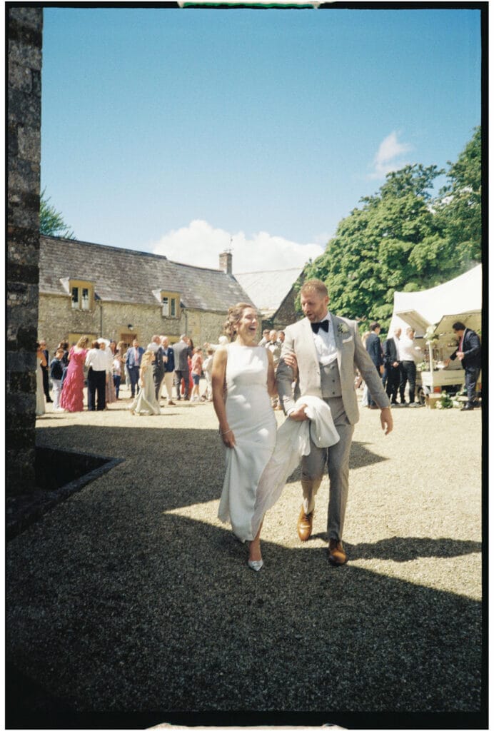 A bride and groom walk together outdoors at a wedding venue, with guests socializing in the background under a blue sky.