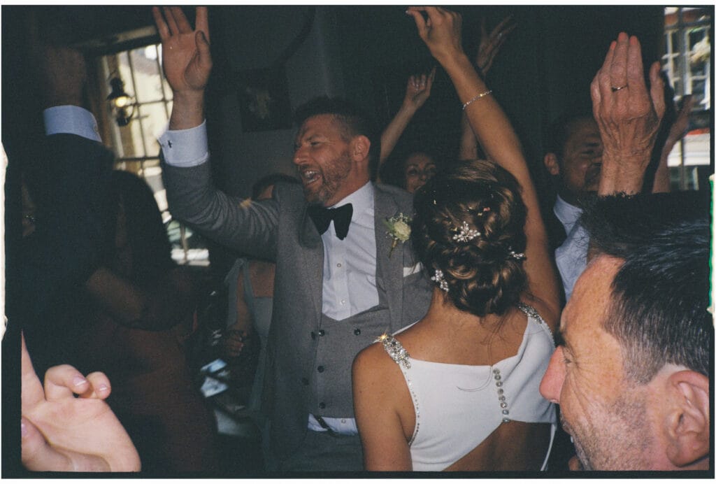 A man in a grey suit and bow tie dances with others at an indoor event; people around him have raised hands and appear to be celebrating.