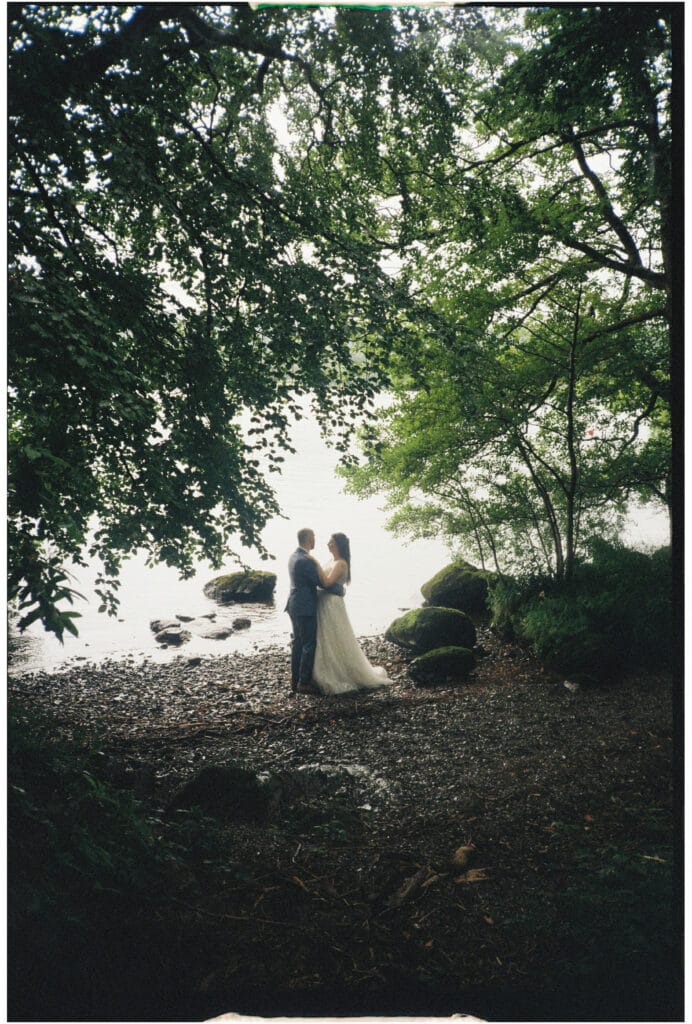 A couple in formal attire stands together by a lake, surrounded by trees and rocks, with water visible in the background.