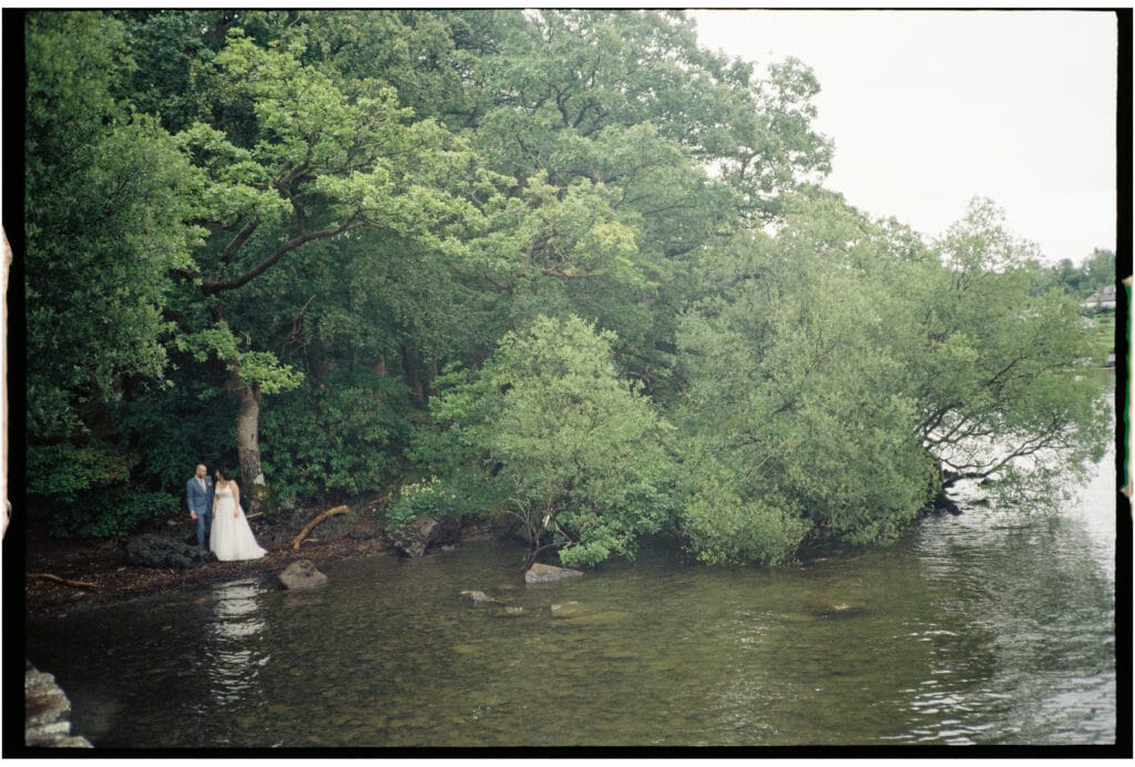 A bride and groom stand at the edge of a wooded shoreline beside calm water, surrounded by dense green trees.