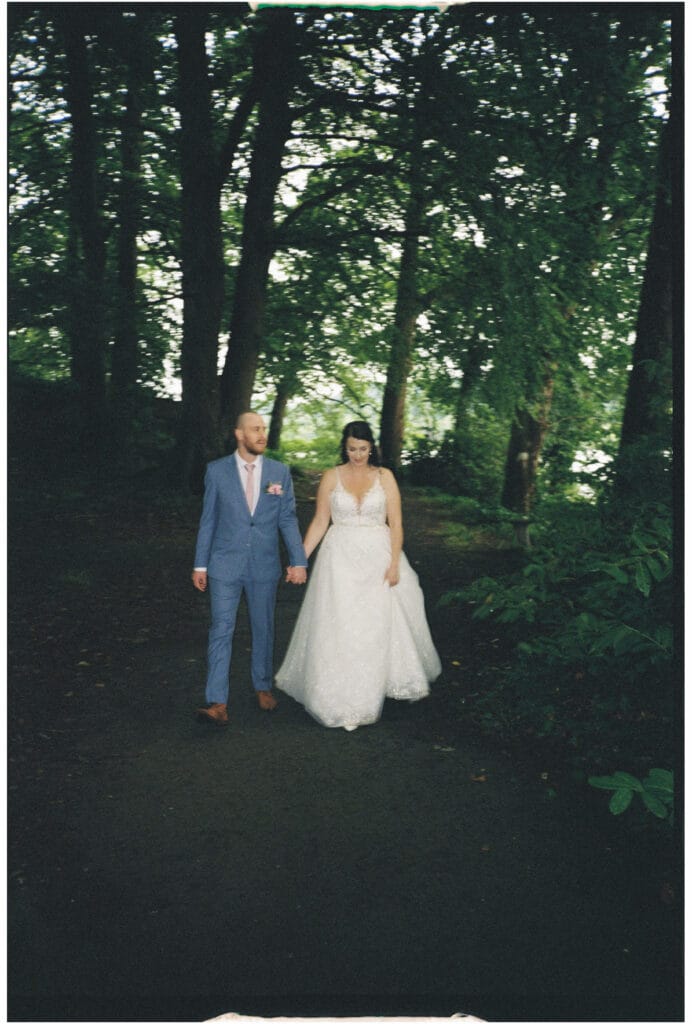 A bride in a white wedding dress and a groom in a blue suit walk hand-in-hand along a shaded path in a wooded area.