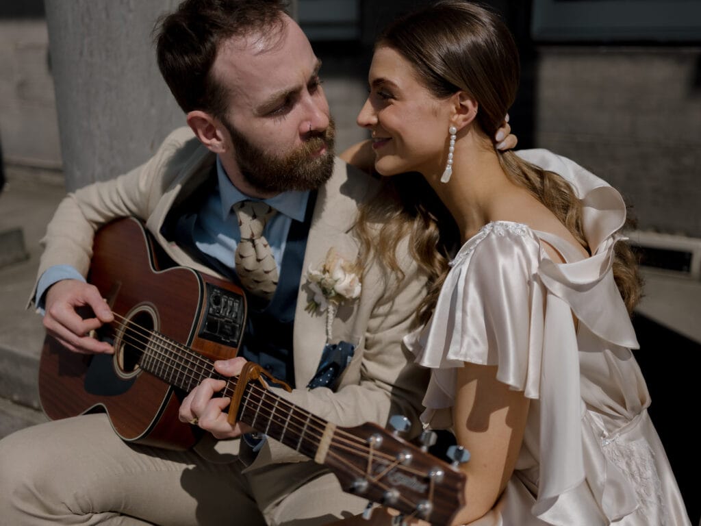 A man in a beige suit plays acoustic guitar while sitting close to a woman in a white dress who is smiling at him.