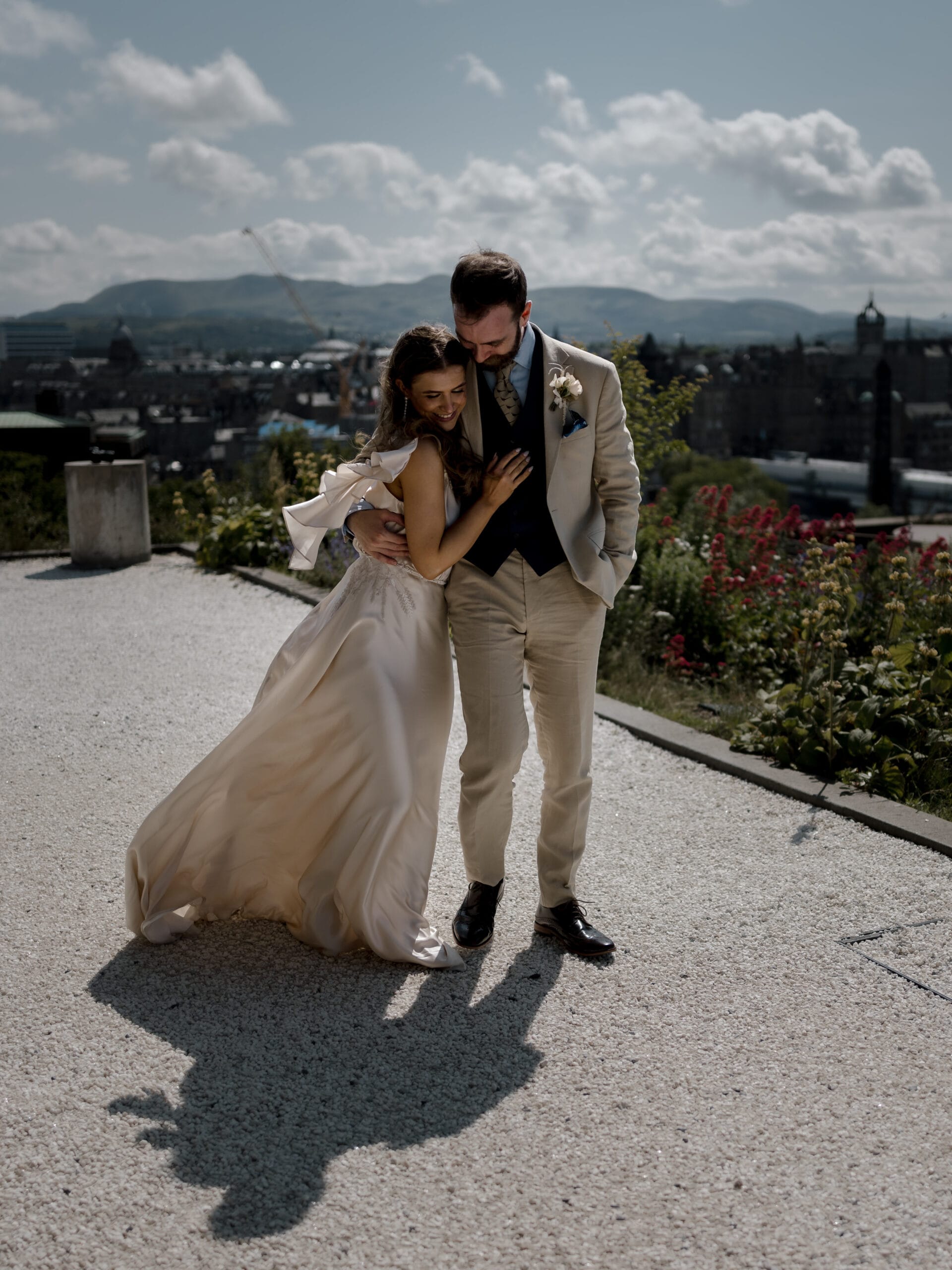 A bride and groom in formal attire embrace outdoors on a sunny day, with city buildings and hills visible in the background.