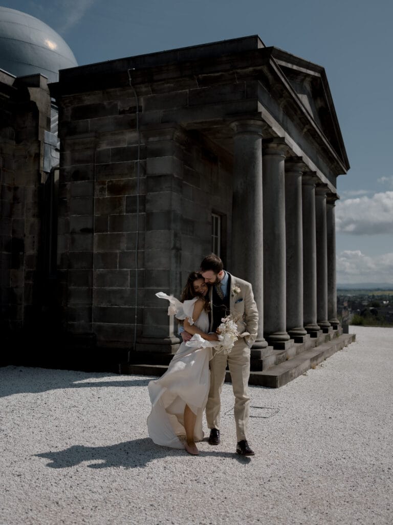 A couple dressed in formal attire stands in front of a stone building with columns on a sunny day. The woman holds a bouquet of flowers while the man kisses her head.