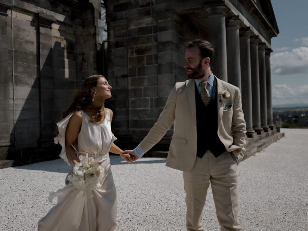 A bride and groom holding hands and smiling at each other, standing outside a stone building with columns on a sunny day.