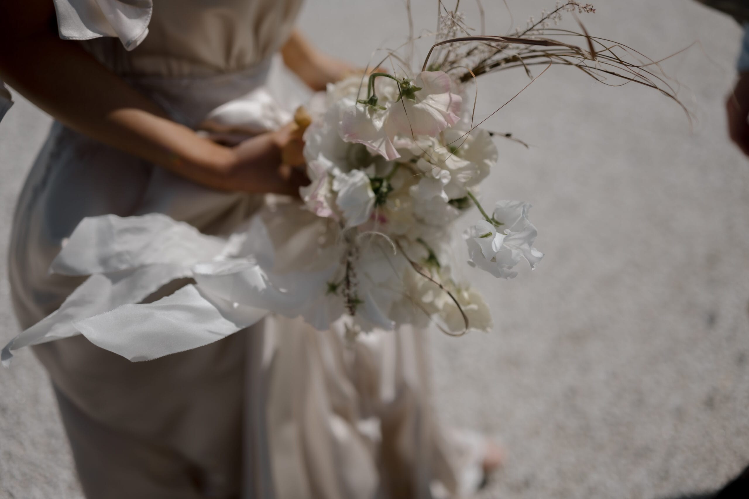 A person in a light-colored dress holds a bouquet of white and pale pink flowers with ribbon, standing on a gravel surface.
