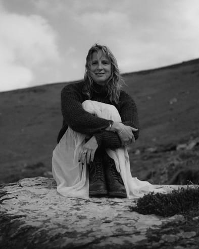 A woman sits on a rock outdoors, wearing a sweater, long skirt, and boots, with hills in the background under a cloudy sky. The image is in black and white.
